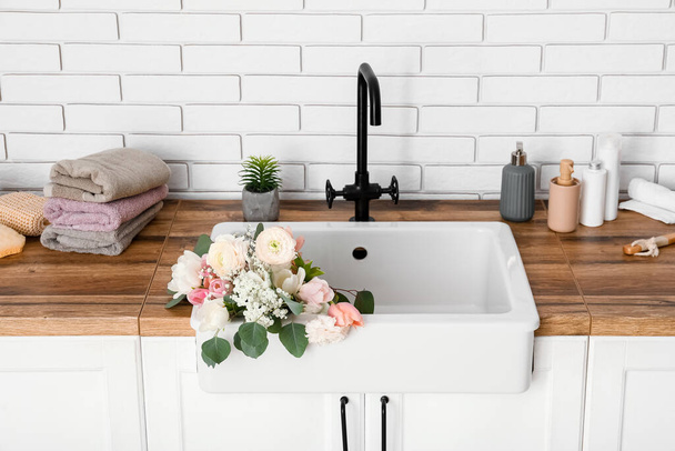 Sink with beautiful flowers and bath supplies on counters near white brick wall - 写真・画像
