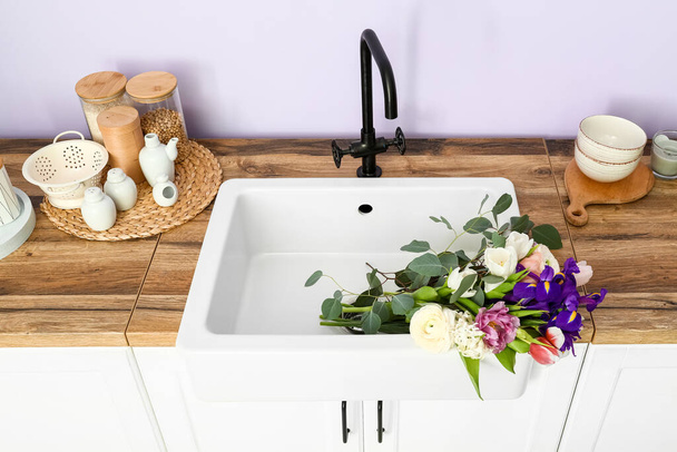 Раковина з красивими квітами та посудом на прилавках біля бузкової стіни
 - Фото, зображення