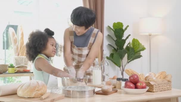 Famiglia afroamericana con madre che indossa grembiule farina trebbia per cucinare e ballare con figlia insieme in cucina a casa, genitore e bambino che preparano cibo con divertimento e giocosità. - Filmati, video