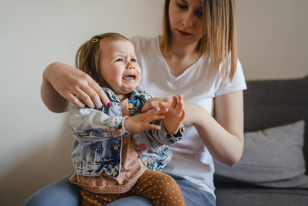 Small caucasian baby girl infant child with her mother dressing undressing small daughter crying unhappy frustrated at home childhood parenthood and growing up family concept real people copy space - Foto, Bild