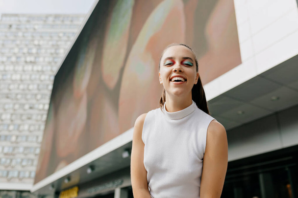 Optimistisch schattig meisje met verzameld haar in wit shirt glimlacht oprecht in de stad. Cool dame in lichte blouse met kijkt in de camera op straat. Hoge kwaliteit foto - Foto, afbeelding