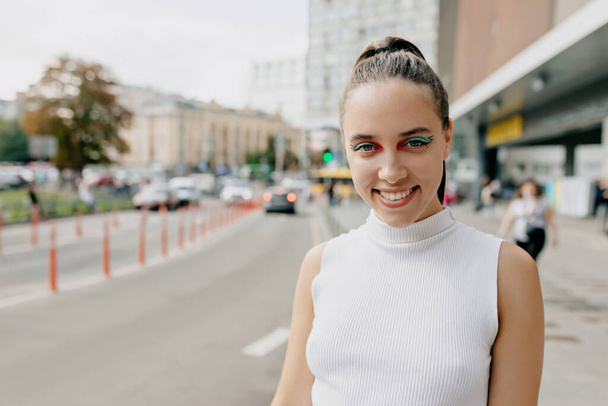 Friendly lovely girl with bright make up and dark collected hair dressed white t-shirt is smiling at camera and spend time outdoor in the city center in warm sunny day. High quality photo - Φωτογραφία, εικόνα