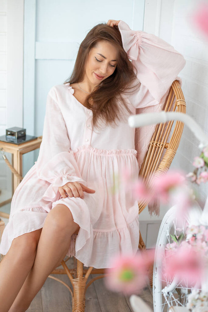 Portrait of young beautiful woman with long hair wearing pink summer dress, sitting on wicker chair near white bicycle. - Photo, Image