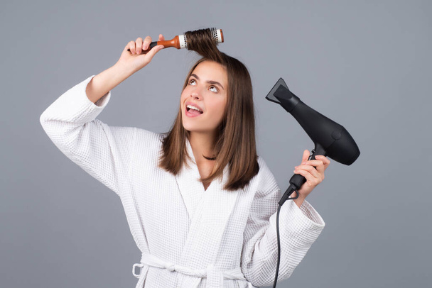Amazed woman in bathrobe dryer hair with hairdryer. Portrait of female model with a comb brushing hair. Girl with hairbrush and hair dryer, hair care and beauty. - Foto, Imagem