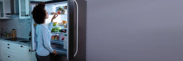 Side View Of Young African Woman Searching Food In An Open Refrigerator - Valokuva, kuva