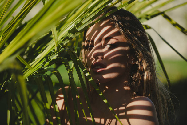 Portrait of young woman behind palm leaves. Exotic tropical leaves near her face. Beauty, fashion, cosmetology, nature and summer spa. - Foto, imagen