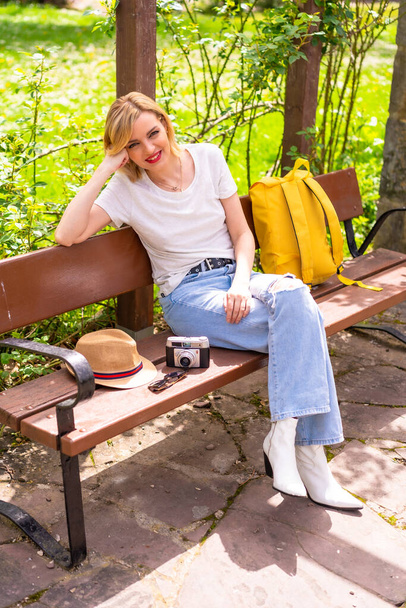 Portrait of a blonde tourist girl on summer vacation sitting on a bench in a park, wearing a hat, photo camera, yellow backpack - Zdjęcie, obraz