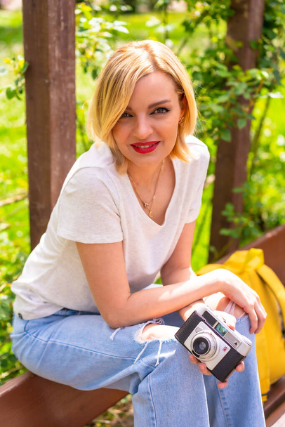 A blonde tourist girl on summer vacation sitting on a bench in a park, wearing a hat, photo camera, yellow backpack - Fotografie, Obrázek