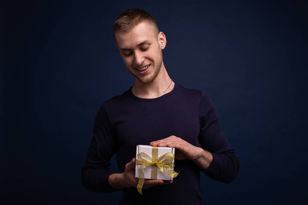 Mavi süveterli, gülümseyen genç bir adam mavi arka planda poz veriyor, elinde bir hediye tutuyor. - Fotoğraf, Görsel