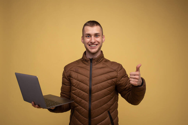 Young 20s man in a brown jacket, on a yellow background, holding a laptop computer, shows a finger at a class, gesture - 写真・画像