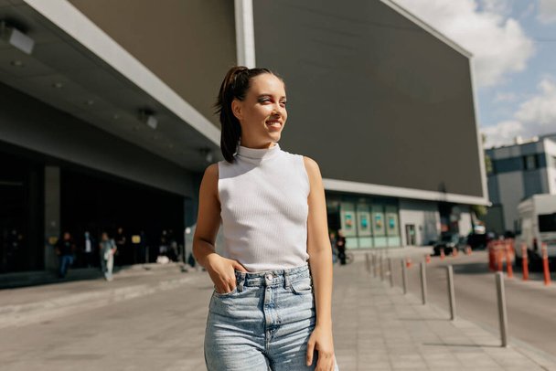 Portrait of young beautiful girl smiling while looking aside hiding from sun outdoors. Long-haired woman with bright make up is wearing white top, denim shorts  - 写真・画像