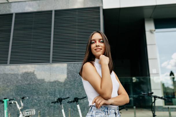 Wonderful stylish young lady with bright make up and dark hair dressed white t-shirt is looking at camera and posing outdoor in city center. High quality photo - Valokuva, kuva