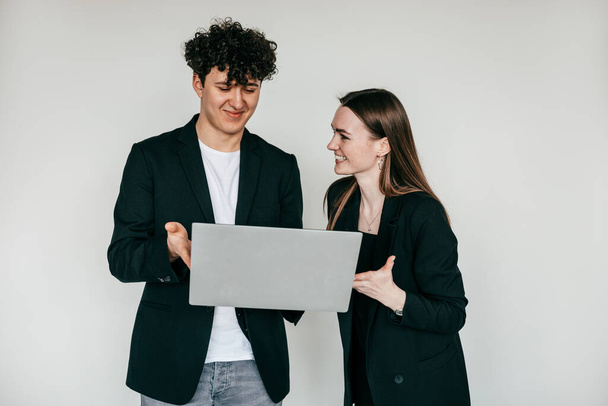 Χαρούμενο ζευγάρι ποζάρει με μοντέρνο laptop και φοράει κομψά μαύρα μπλέιζερ και αξεσουάρ για άνδρες και γυναίκες, λευκό φόντο. Κατάλογος στυλ μόδας ρούχων για επιχειρηματίες, ηλεκτρονικά - Φωτογραφία, εικόνα
