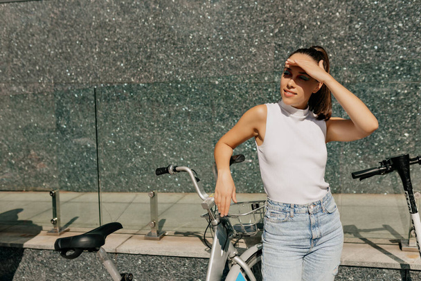 Pretty trendy girl with collected dark hair wearing white t-shirt and jeans is covering face from sun and looking aside while standing near the bike . High quality photo - Foto, immagini
