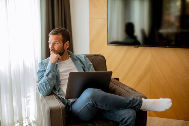 Красивый молодой человек, использующий ноутбук в гостиной - Фото, изображение