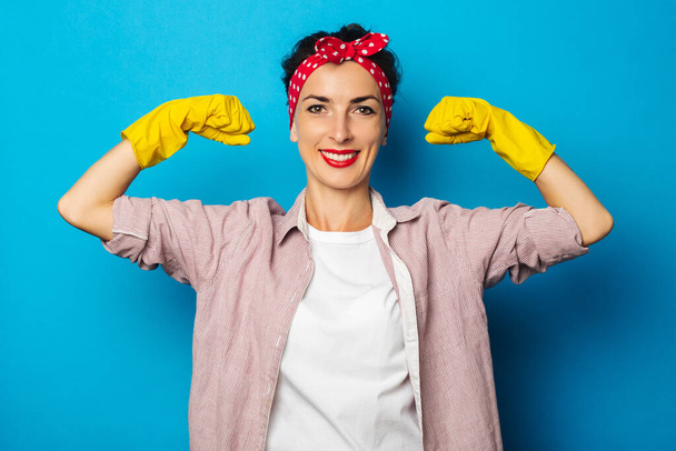 Smiling young woman showing biceps in cleaning gloves on blue background - Photo, Image