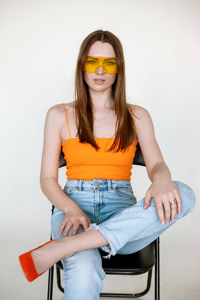 Hot woman with brown hair and stylish sunglasses sitting on black chair and wearing seasonal blue denim and bright orange top, white background. Fashion catalog of casual clothes for young ladies - Photo, Image