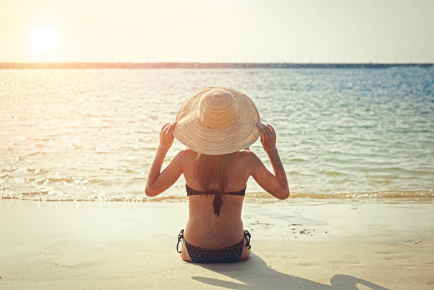 Young girl on the beach sits with her back to the sea in a hat - Фото, изображение