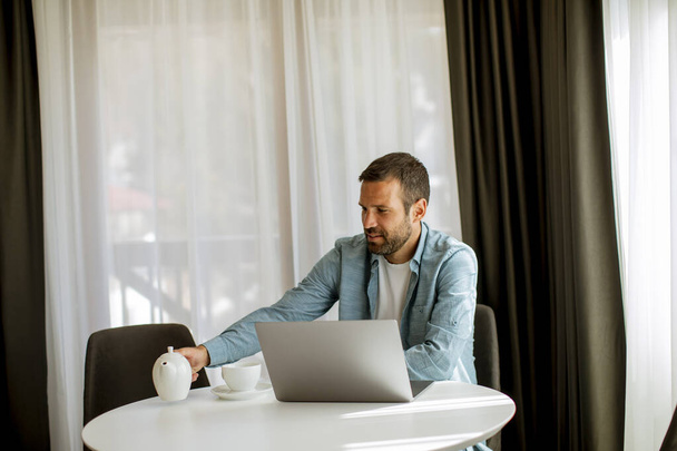Hombre joven guapo usando el ordenador portátil y beber té en la sala de estar - Foto, Imagen