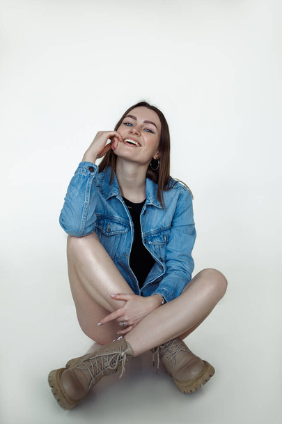 Smiling hot woman with brown hair and perfect makeup posing and wearing seasonal blue denim jacket and leather boots, white background. Fashion style catalog of casual clothes for young ladies - Фото, зображення