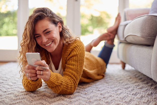 Woman In Warm Jumper Lying On Floor At Home Using Mobile Phone - Photo, image