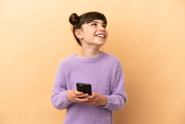Petite fille caucasienne isolée sur fond beige en utilisant le téléphone mobile et en regardant vers le haut - Photo, image
