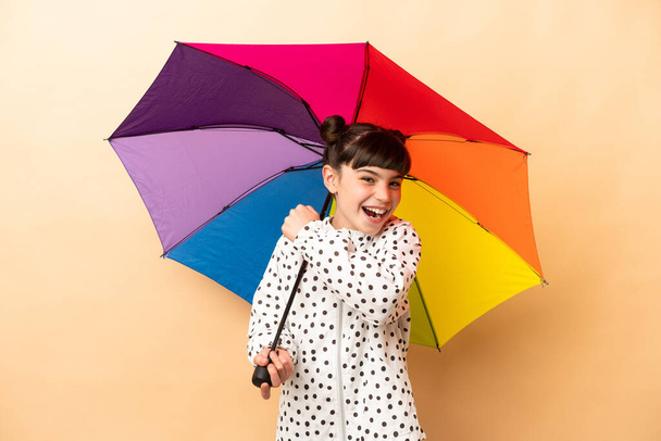 Little girl holding an umbrella isolated on beige background celebrating a victory - Photo, image