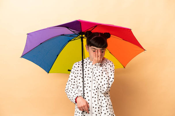 Little girl holding an umbrella isolated on beige background with tired and sick expression - Foto, Imagen