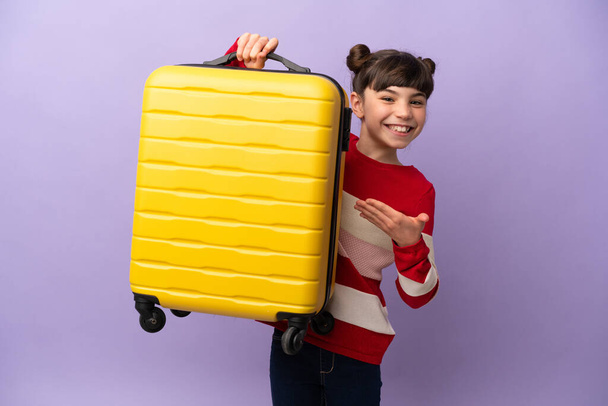 Little caucasian girl isolated on purple background in vacation with travel suitcase - Photo, Image