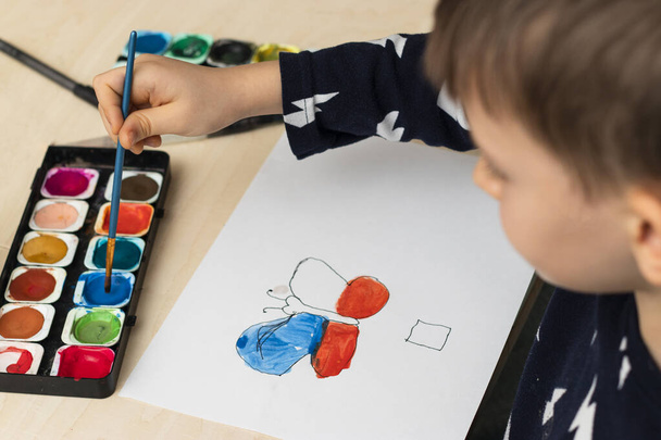 Χαριτωμένο αγόρι κάνει τα μαθήματά του, χρωματίζει σελίδες, γράφει και ζωγραφίζει. Τα παιδιά ζωγραφίζουν. Δημιουργικό αγόρι. ζωγραφική χεριών, κορυφαία προβολή - Φωτογραφία, εικόνα