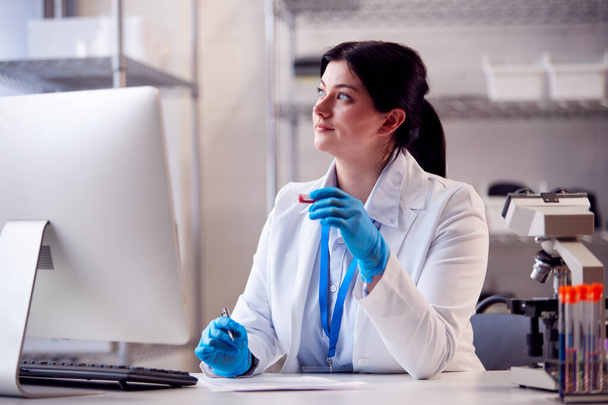 Female Lab Worker Wearing PPE Face Shield Recording Blood Test Results On Computer - Foto, Imagen