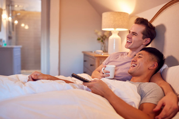 Loving Same Sex Male Couple Lying In Bed At Home Watching TV Together - Фото, изображение