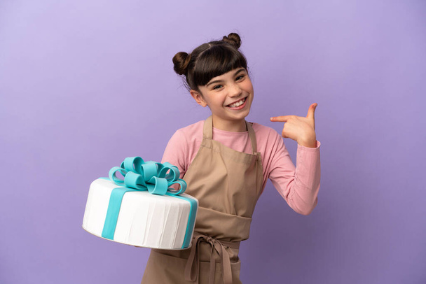 Pastry little girl holding a big cake isolated on purple background giving a thumbs up gesture - Zdjęcie, obraz