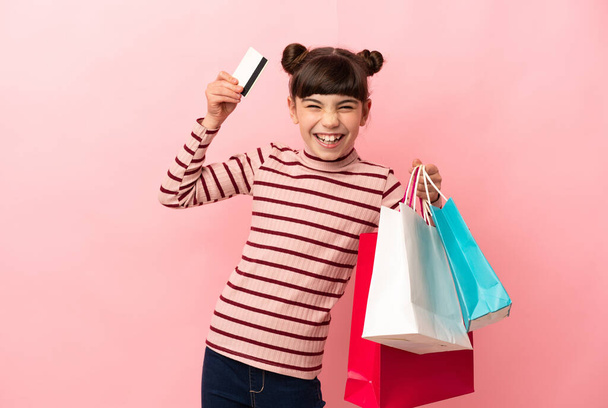 Malá kavkazská dívka izolované na růžovém pozadí držení nákupních tašek a kreditní karty - Fotografie, Obrázek