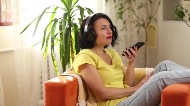 Γυναίκα σε hedphones, lyuing στον καναπέ, χρήση εικονικών εντολών βοηθός στο smartphone, φωνητική μεταφραστής έννοια διαφήμισης εφαρμογή, νέα έννοια της τεχνολογίας, θηλυκό ενεργοποιούν ψηφιακή ακουστικό βοηθό στο gadget - Πλάνα, βίντεο