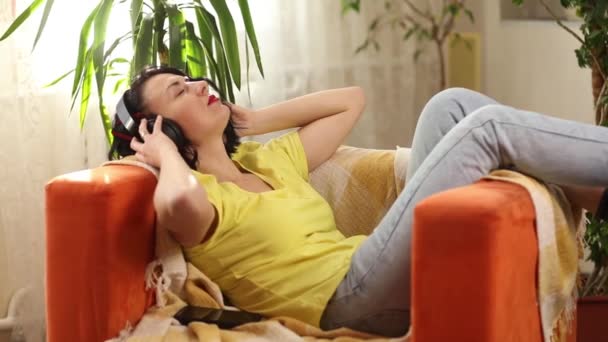 Női nő fülhallgatóban csukott szemmel zenét hallgat, podcast ül egy narancssárga kanapén. Gyönyörű nő fényes ruhában élvezi a zenét otthon - Felvétel, videó