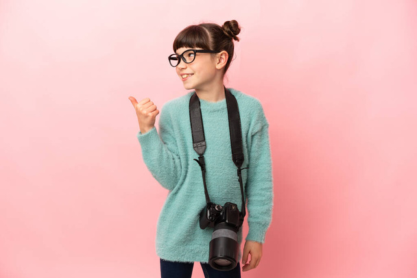 Kleines Fotografenmädchen auf rosa Hintergrund, das zur Seite zeigt, um ein Produkt zu präsentieren - Foto, Bild