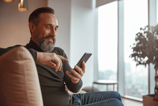Ευτυχισμένος γενειοφόρος άνδρας κάθεται και πληκτρολογώντας μήνυμα στο κινητό τηλέφωνο - Φωτογραφία, εικόνα