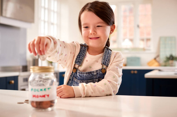 Girl Saving Money Into Jar Labelled Pocket Money On Kitchen Counter At Home - Foto, afbeelding