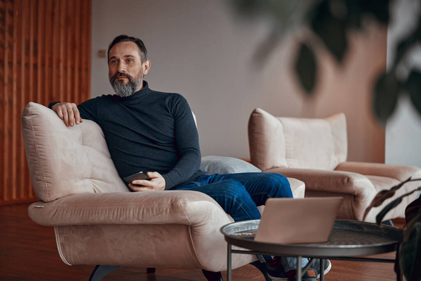 Взрослый мужчина с бородой сидит в кресле и держит телефон - Фото, изображение