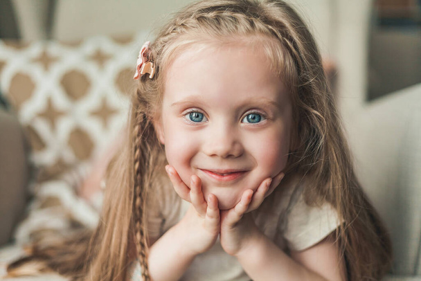 Una niña linda de ojos azules de 4 años con el pelo largo es feliz sentado en un sillón en una casa acogedora. - Foto, Imagen