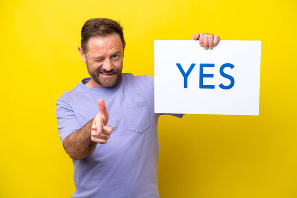 Middle age caucasian man isolated on yellow background holding a placard with text YES and pointing to the front - Photo, image