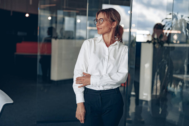 Focused business woman wearing eyeaglasses looks away during the working day at modern office - Zdjęcie, obraz