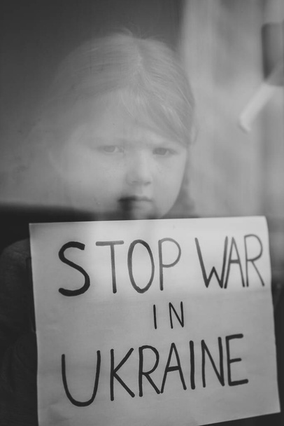 Child at the window and calls No War. Poor girl protesting war conflict raises banner with inscription Stop war in Ukraine. Stop russian aggression. Evacuation of civilians. Black and white photo. - Foto, afbeelding