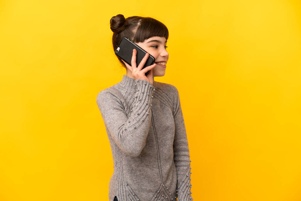 Little caucasian girl isolated on yellow background keeping a conversation with the mobile phone with someone - Foto, Imagen