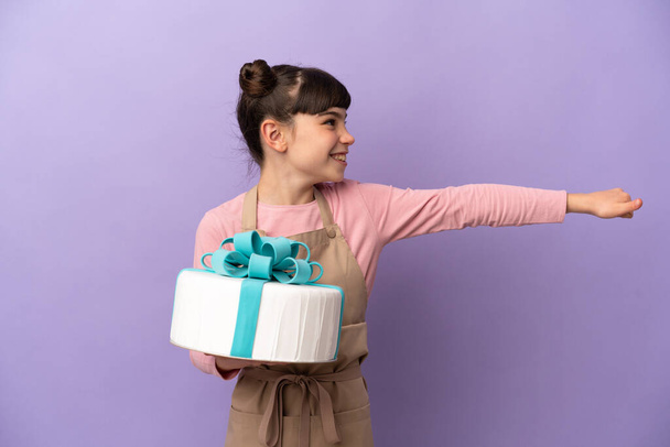 Gebäck kleines Mädchen hält einen großen Kuchen isoliert auf lila Hintergrund gibt einen Daumen nach oben Geste - Foto, Bild