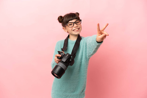 Kleines Fotografenmädchen auf rosa Hintergrund lächelt und zeigt Siegeszeichen - Foto, Bild