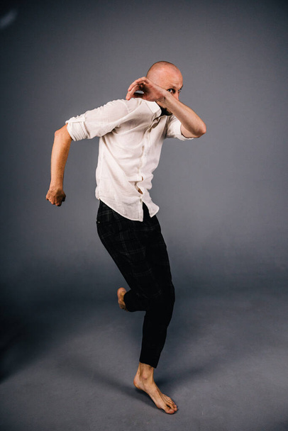 studio portrait of a bald guy with a beautiful beard barefoot in a white shirt and plaid pants dancing on a gray background - Photo, image