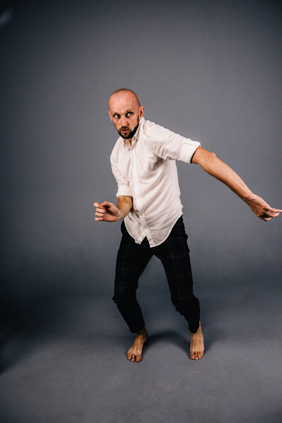 studio portrait of a bald guy with a beautiful beard barefoot in a white shirt and plaid pants dancing on a gray background - Zdjęcie, obraz