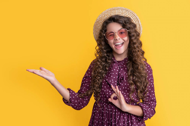 happy child in straw hat and sunglasses with long brunette curly hair presenting product - Foto, Imagen